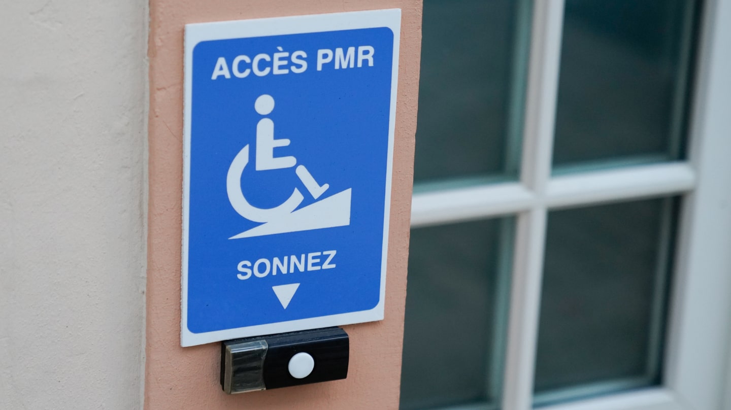 Un panneau bleu indiquant l'accès PMR avec une représentation de personne en fauteuil roulant et une sonnette.