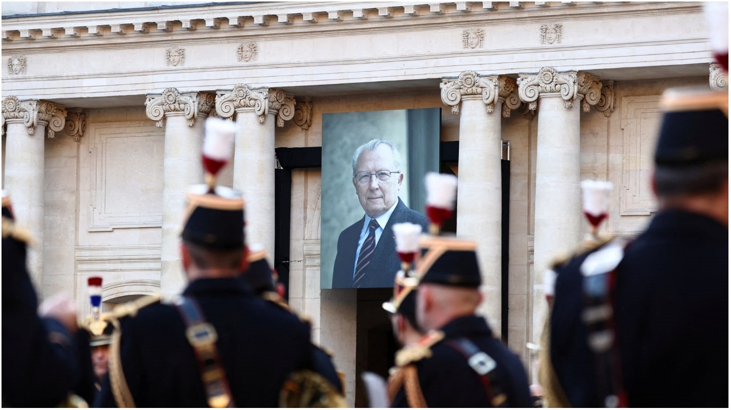 Un grand portrait de Jacques Delors est accroché dans la cour des Invalides ; des gardes au premier plan.