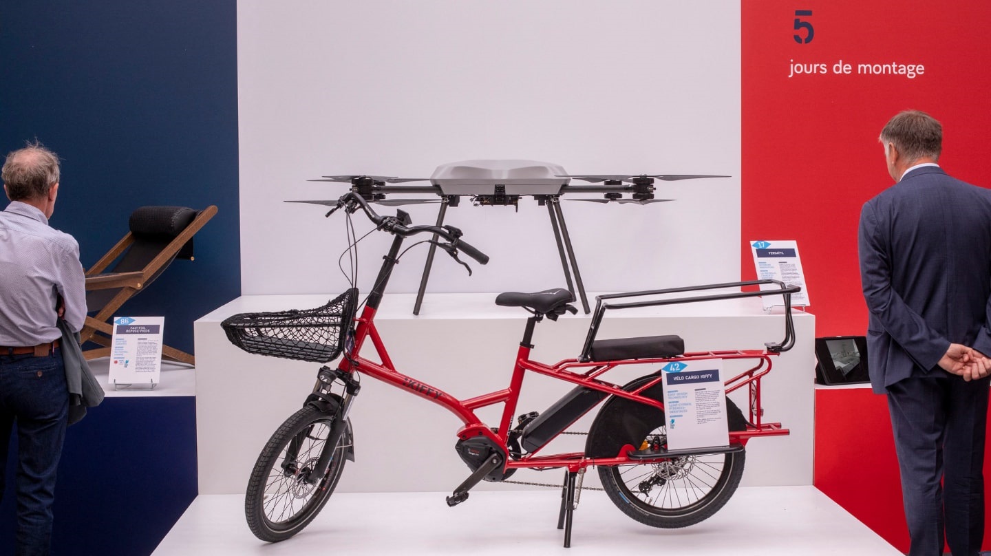 Un vélo rouge au premier plan, un drone devant un fond tricolore, deux hommes de dos qui regardent d'autres objets exposés.