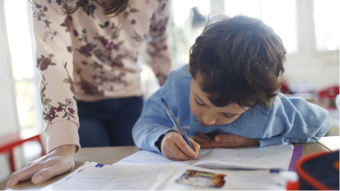 Enfant en train d'écrire, supervisé par une femme.